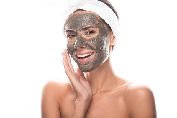 Nude young woman in cosmetic hair band with clay mask on face looking at camera with smile isolated on white — Stock Photo