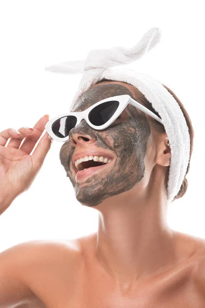 Nudo sorridente giovane donna in fascia di capelli cosmetici e occhiali da sole con maschera di argilla sul viso isolato su bianco — Foto stock