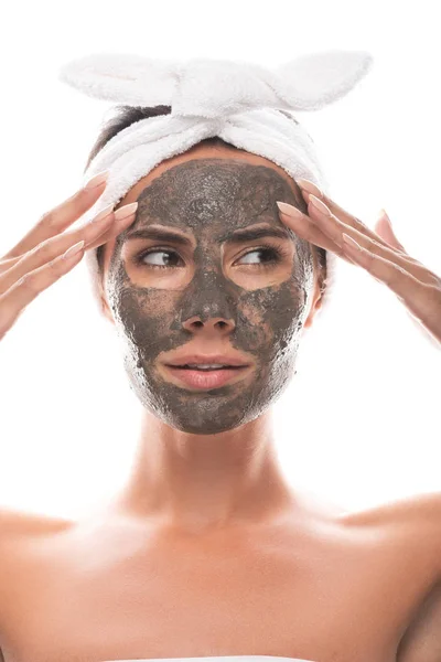 Front view of pensive nude young woman in cosmetic hair band with clay mask on face isolated on white — Stock Photo