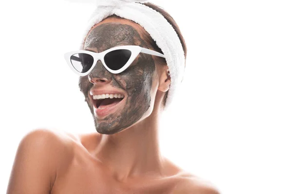 Обнаженная молодая женщина в косметическом браслете и солнцезащитных очках с глиняной маской на лице, смеющаяся изолированно на белом — стоковое фото