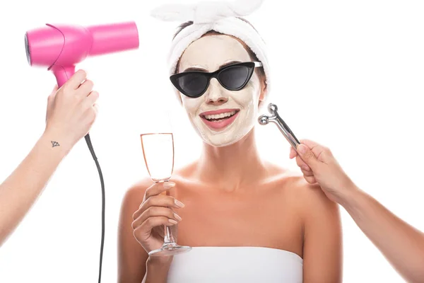Vista parziale di estetiste in possesso di asciugacapelli e massaggiatore facciale e donna sorridente in occhiali da sole con maschera facciale in possesso di bicchiere di vino isolato su bianco — Foto stock