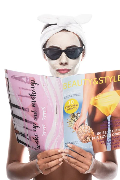 Frontansicht einer Frau mit kosmetischem Haarband und Sonnenbrille mit Gesichtsmaske, die Zeitschrift auf weiß isoliert liest — Stockfoto