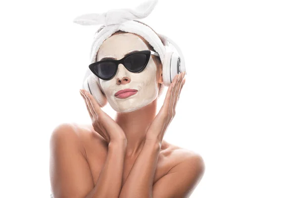 Mujer desnuda en banda cosmética para el cabello y gafas de sol con máscara facial escuchando música en auriculares aislados en blanco - foto de stock