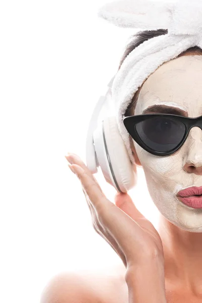 Vista recortada de mujer desnuda en banda de pelo cosmético y gafas de sol con máscara facial escuchando música en auriculares aislados en blanco - foto de stock