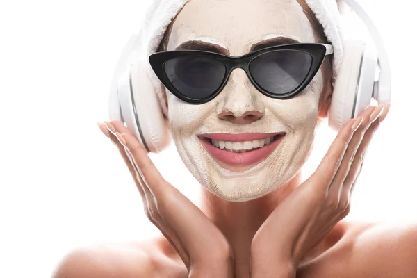 Вид обнаженной женщины в косметической повязке для волос и солнцезащитных очках с маской для лица, слушающей музыку в наушниках, изолированных на белом фоне — стоковое фото
