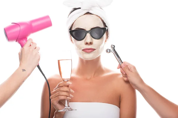 Vue partielle des esthéticiennes tenant le sèche-cheveux et le masseur facial et de la femme dans des lunettes de soleil avec masque facial tenant le verre à vin isolé sur blanc — Photo de stock