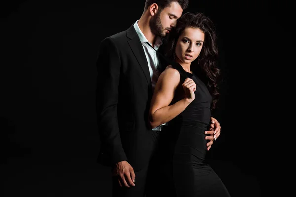 Hombre con los ojos cerrados abrazando hermosa mujer en vestido mirando a la cámara aislada en negro - foto de stock