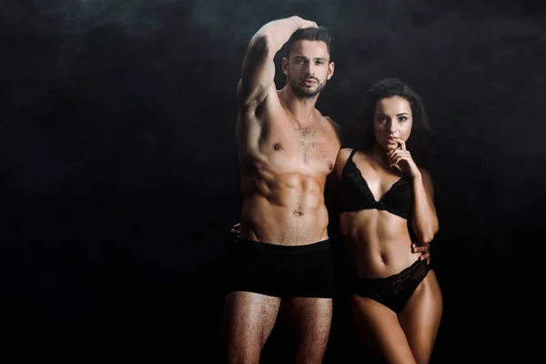 Handsome shirtless man standing and touching hair near attractive girl in lingerie on black with smoke — Stock Photo