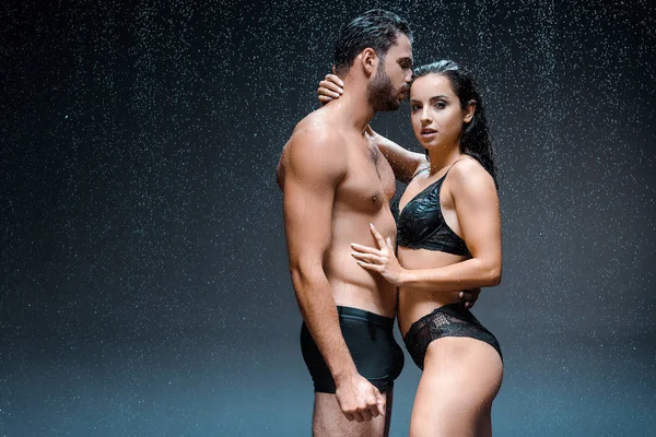 Sexy hombre sin camisa de pie con mujer joven en lencería bajo gotas de lluvia en negro - foto de stock