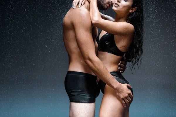 Обрізаний вид сексуального чоловіка обіймає і цілує мокру жінку в білизні під дощами на чорному — стокове фото