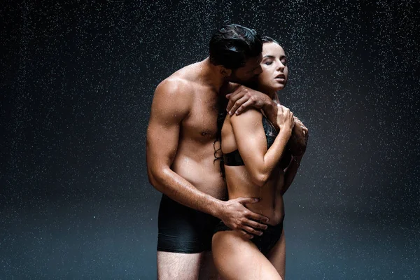 Homem sexy abraçando e beijando mulher molhada em roupa interior sob gotas de chuva em preto — Fotografia de Stock