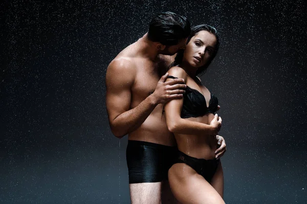 Shirtless man hugging and kissing wet woman in underwear under raindrops on black — Stock Photo