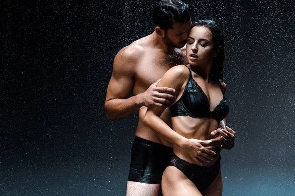 Shirtless man hugging sexy and wet girl under raindrops on black — Stock Photo