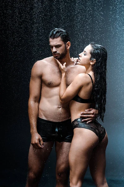 Сексуальная и мокрая девушка, касающаяся лица мужчины без рубашки, стоящего под каплями дождя на черном — стоковое фото
