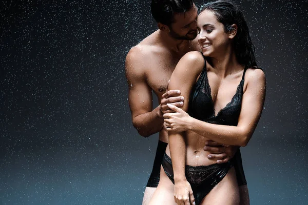 Shirtless hombre abrazando feliz mojado mujer bajo gotas de lluvia en negro - foto de stock