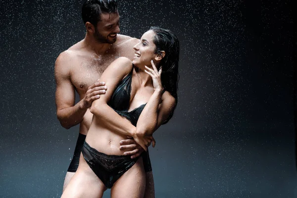 Feliz musculoso hombre abrazando alegre mojado mujer bajo gotas de lluvia en negro - foto de stock