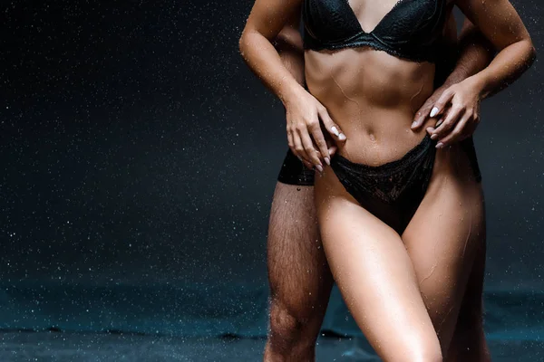 Cropped view of muscular man hugging young wet woman under raindrops on black — Stock Photo