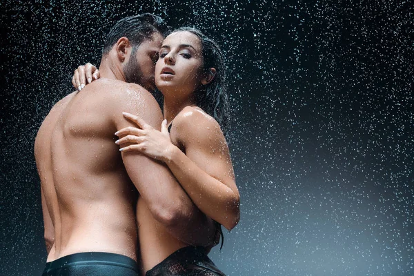 Sexy barbudo hombre abrazando apasionado chica bajo gotas de lluvia en negro - foto de stock