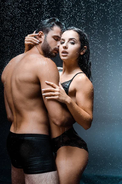Bonito homem abraçando molhado e sexy namorada sob raindrops no preto — Fotografia de Stock
