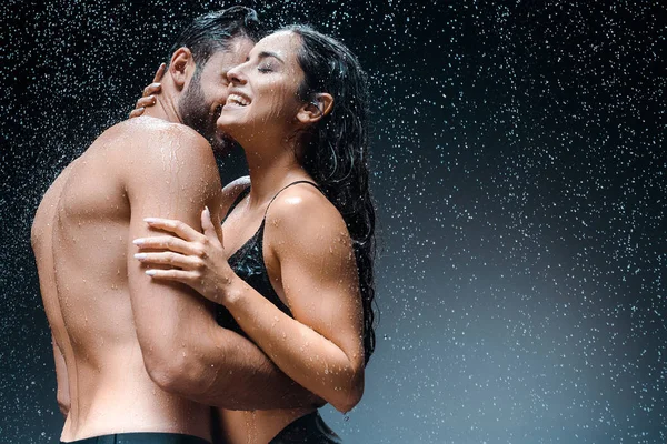 Húmedo sin camisa hombre abrazando feliz novia bajo gotas de lluvia en negro - foto de stock