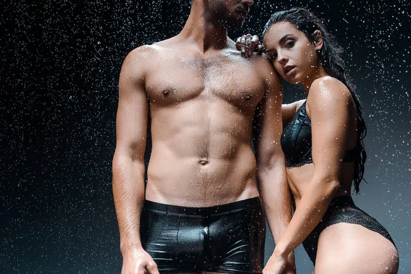 Corte vista de molhado sem camisa homem de pé com namorada sob gotas de chuva em preto — Fotografia de Stock