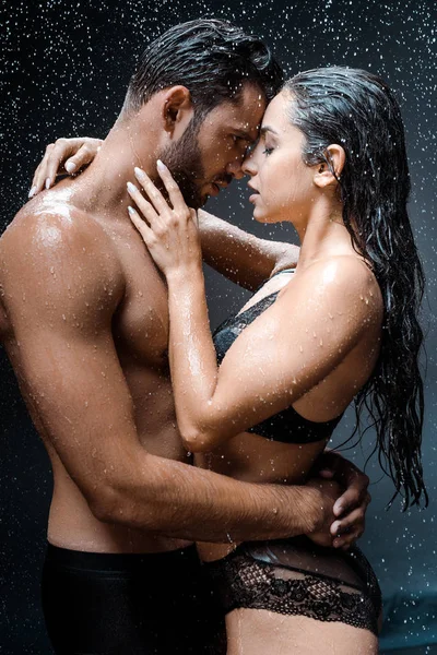 Attractive woman touching face of shirtless and wet man under raindrops on black — Stock Photo