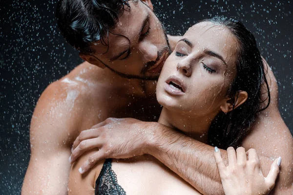 Uomo senza maglietta e bagnato abbracciare fidanzata attraente con gli occhi chiusi sotto gocce di pioggia sul nero — Foto stock