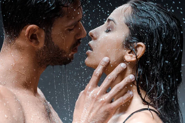 Hombre sin camisa y mojado tocando la cara de novia atractiva con los ojos cerrados bajo gotas de lluvia en negro - foto de stock