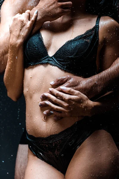 Cropped view of man touching sexy and wet girlfriend under raindrops on black — Stock Photo