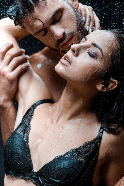 Selective focus of sexy man touching wet girlfriend with closed eyes under raindrops on black — Stock Photo