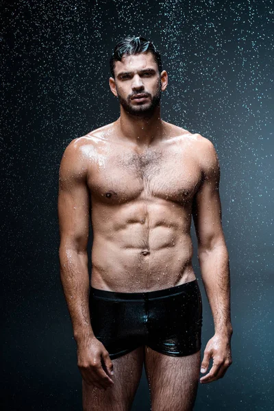 Mojado guapo hombre mirando a cámara mientras de pie bajo gotas de lluvia en negro - foto de stock