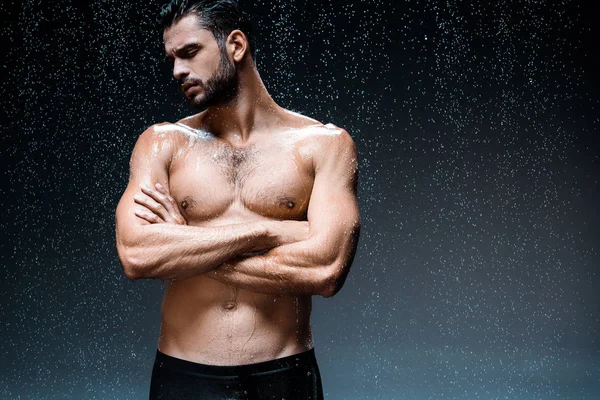 Bell'uomo bagnato in piedi con le braccia incrociate sotto gocce di pioggia sul nero — Foto stock