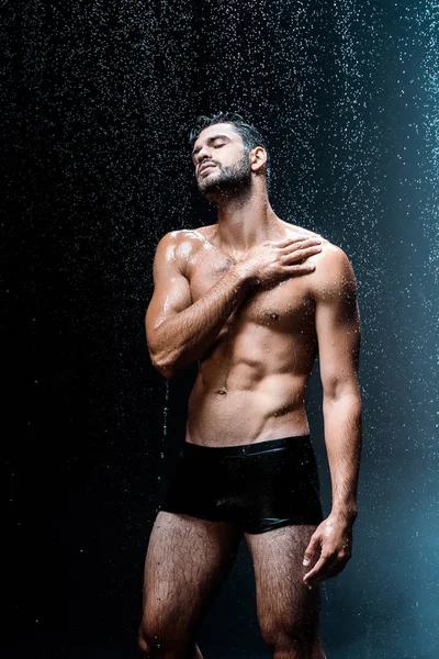 Shirtless and wet man standing under raindrops on black — Stock Photo