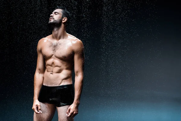 Shirtless and wet man with closed eyes standing under raindrops on black — Stock Photo