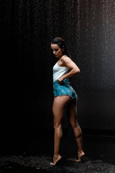 Sexy joven y húmeda mujer de pie en punta del pie bajo gotas de lluvia en negro - foto de stock