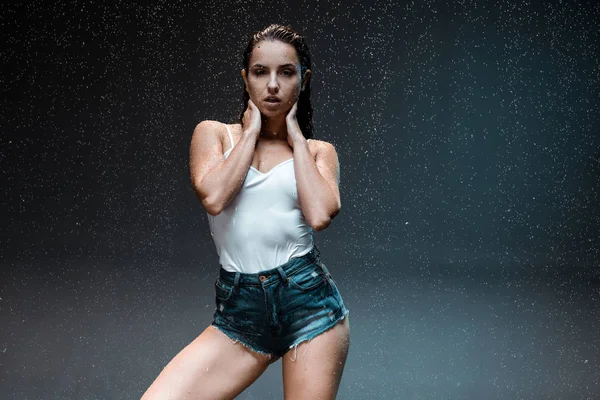 Sexy jovem mulher de pé e olhando para a câmera sob raindrops no preto — Fotografia de Stock