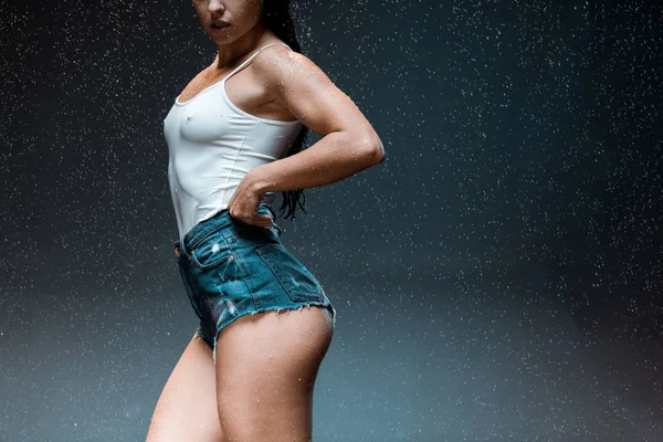 Cropped view of young sexy woman in denim shorts standing under raindrops on black — Stock Photo