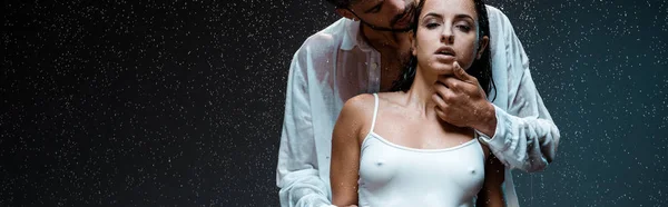 Colpo panoramico di uomo barbuto toccare il viso sulla ragazza sexy sotto gocce di pioggia sul nero — Foto stock