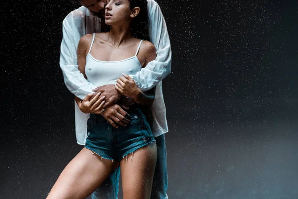 Cropped view of man hugging sexy wet girlfriend under raindrops on black — Stock Photo