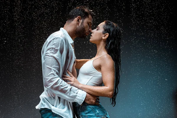 Vue latérale de bel homme embrassant sexy copine humide sous gouttes de pluie sur noir — Photo de stock