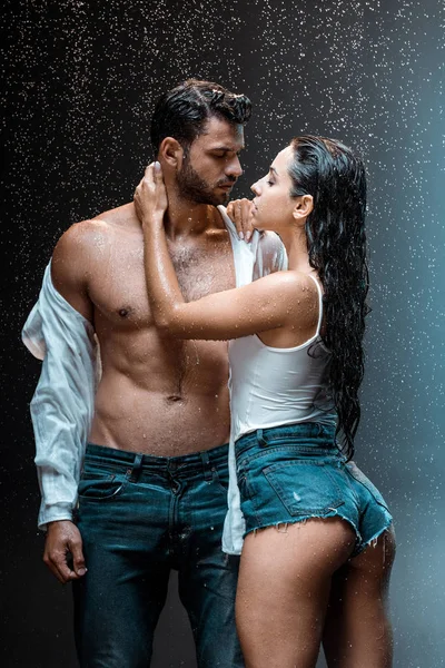 Déshabillé et musclé homme debout avec sexy copine sous gouttes de pluie sur noir — Photo de stock