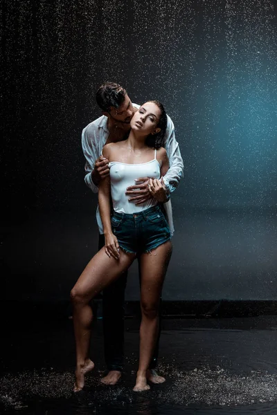 Handsome man standing and kissing sexy girlfriend under raindrops on black — Stock Photo