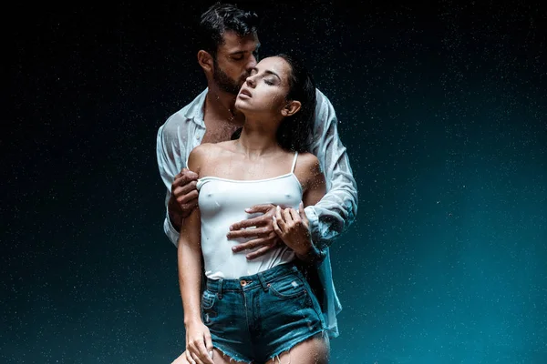 Handsome man standing and kissing sexy girlfriend with closed eyes under raindrops on black — Stock Photo