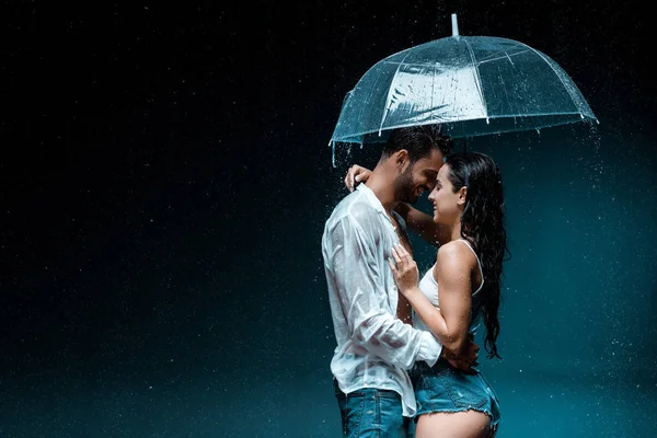 Side view of cheerful girl standing with handsome man and umbrella on black — Stock Photo