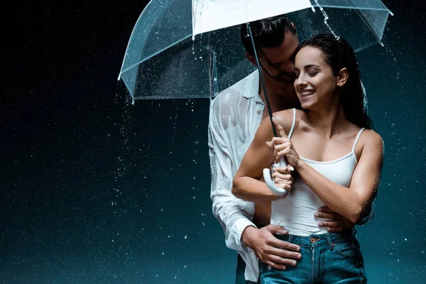 Alegre chica sosteniendo paraguas y de pie con guapo novio bajo gotas de lluvia en negro - foto de stock