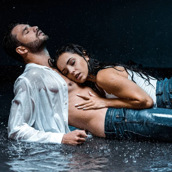 Sexy and wet girl lying with handsome boyfriend under raindrops on black — Stock Photo