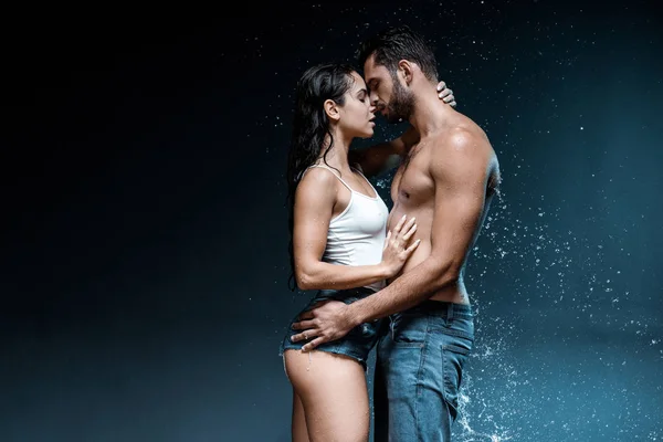 Sexy sin camisa hombre abrazando atractiva mujer cerca de salpicadura de agua en negro - foto de stock