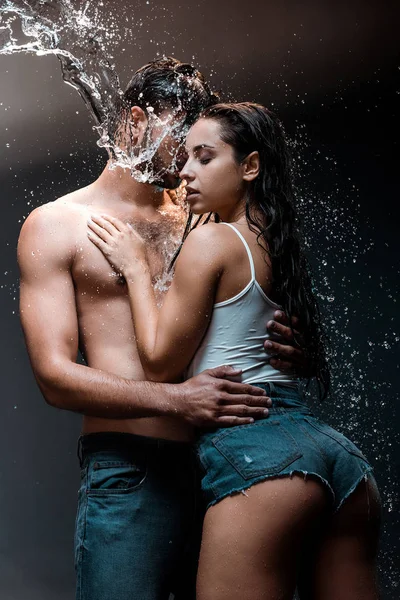 Shirtless man embracing girlfriend in denim shorts near splash of water on black — Stock Photo