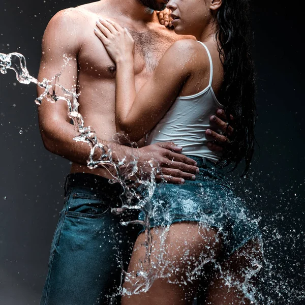 Обрезанный вид мужчины без рубашки, обнимающего девушку в джинсовых шортах возле брызг воды на черном — стоковое фото