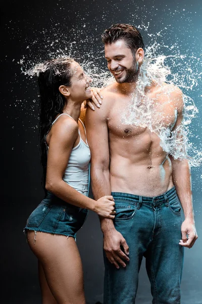 Hombre sin camisa sonriendo con atractiva novia en pantalones cortos de mezclilla cerca de salpicadura de agua en negro - foto de stock